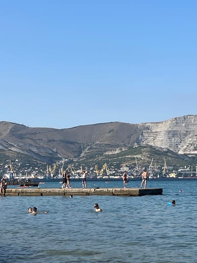 The image depicts a sunny day at a coastal area, likely in Ol'ginskii, Russia, given the context. The scene is dominated by a vast expanse of blue water on the right and a clear blue sky on the left. In the background, a steep, rocky mountain looms over a coastal town with buildings and numerous yellow cranes, suggesting a port or industrial area.

Along a concrete pier extending into the water, several people are visible. Some are standing on the pier, appearing to be enjoying the view or preparing to enter the water. Others are in the water, swimming or wading. The water is calm with gentle ripples. The overall mood appears relaxed and recreational, typical of a summer day at the beach. The time of day is clearly daytime, indicated by the bright sunlight casting shadows. No visible text is present in the image.