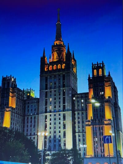 This image depicts the Kotelnicheskaya Embankment Building in Moscow, Russia, at twilight. The monumental Stalinist skyscraper, characterized by its imposing architecture and prominent spire topped with a star, is illuminated by warm yellow lights highlighting its decorative elements and numerous windows. The building is framed by a deep blue sky, suggesting the late evening or early morning. In the foreground, trees partially obscure the base of the building, and streetlights cast a glow on the scene. A blue directional sign with white arrows indicating a split in the road is visible on the right side of the image. There are no people or discernible activity within the frame.