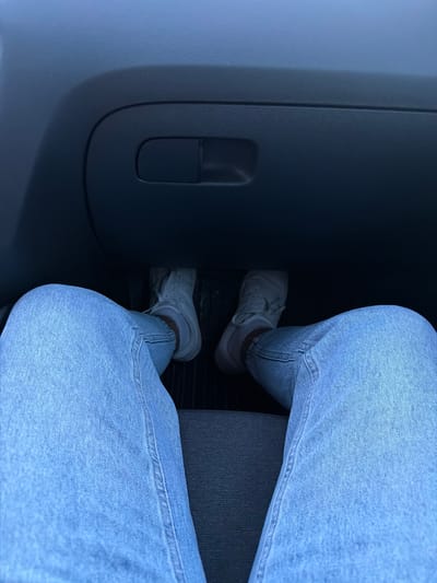 The image shows a person's legs and feet inside a car. The person is wearing light blue jeans and white sneakers. The feet are positioned over what appear to be car pedals. In the background, there is a black dashboard with a glove compartment. The lighting suggests it is daytime. This image does not provide any information to determine the location in the city of Bochnia, Poland.