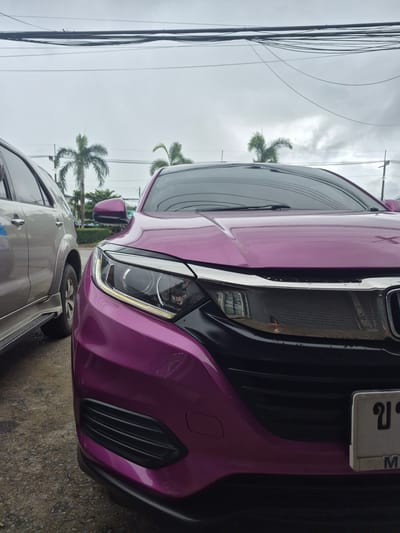This is a close-up, eye-level, outdoor shot of the front of a vibrant magenta-colored car. The car's headlights and grille are prominently featured, with a sliver of a silver SUV visible to its left. The car's license plate, with Thai characters and the letter "M", is partially obscured. In the background, palm trees and utility lines are visible against an overcast sky. The scene appears to be on a street in the city of Ban Yid, Thailand.