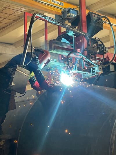 This image captures an industrial welding operation taking place indoors at Tomba, Montenegro. A welder, clad in full protective gear including a red-orange welding helmet, dark jacket, and protective gloves, is actively engaged in welding a large, dark metal cylindrical structure. Bright, intense white and blue sparks, characteristic of arc welding, erupt from the point of contact, illuminating the immediate work area and casting a strong glow.
The scene is set within a large industrial facility, indicated by the presence of heavy machinery and structural components. Above the welder, a robust gantry or crane system is visible, featuring both yellow and red beams. On a prominent yellow beam, the text "ABUS 5t" is clearly legible, denoting a brand (ABUS) and a lifting capacity of 5 tons, common for industrial overhead cranes. Various components of the welding system, including a wire feeder and associated cables and hoses, are mounted on the red gantry structure, suggesting a mechanized or semi-automated welding setup assisting the worker. The overall ambiance is industrial, with artificial lighting accentuating the bright welding arc.