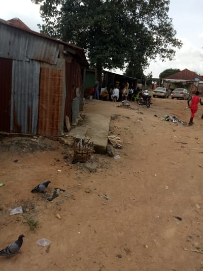 The image depicts a dusty, outdoor scene in Karshi, Nigeria. A corrugated metal structure, appearing weathered and aged, dominates the left side of the frame. In the background, a group of people are gathered, some seated on chairs and others standing. Two cars are parked further in the background, and a motorcycle is visible near the gathering. To the right of the scene, there is a pile of refuse. The ground is covered in reddish-brown soil, and several pigeons are present in the foreground and midground. The overall impression is one of a rural or semi-urban setting with signs of daily life and some environmental neglect.