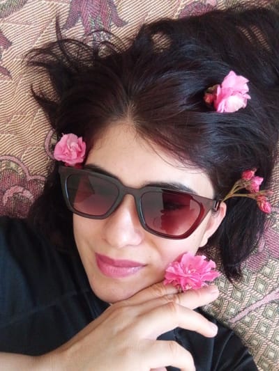 This is a close-up, overhead shot of a woman lying on a patterned textile. She has dark hair adorned with several small pink flowers. The woman is wearing dark-rimmed sunglasses with a reddish tint and has pink lipstick on. Her left hand is resting near her face, with a pink flower placed on her ring finger. She appears to be relaxed, with a gentle smile. The background textile has a diamond pattern in shades of brown, pink, and green. The lighting suggests it may be daytime, possibly indoors given the lack of strong shadows, or outdoors on a bright, overcast day. There is no visible text in the image. The setting is likely domestic, given the textile and relaxed pose.
