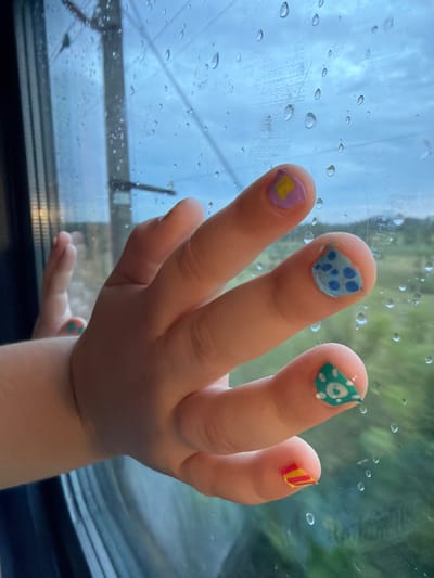 A close-up shot shows a child's hand with decorated fingernails against a window pane covered in raindrops. The child's fingers are extended towards the window, with each nail adorned with a different design. The thumb has a light purple base with a yellow diamond shape. The index finger features a light blue base with darker blue polka dots. The middle finger has a light green base with white circular patterns. The ring finger displays horizontal stripes of red and yellow. Outside the window, a blurred green landscape is visible, suggesting the hand is inside a moving vehicle, possibly a train or car, as indicated by the vertical lines and structure visible through the glass. The sky appears overcast with diffused light. The location is not identifiable from the visual information provided in the image.