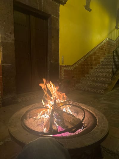 A stone fire pit, complete with a wire mesh dome, hosts a vibrant fire. Flames lick outward, illuminating the immediate surroundings. In the background, a mustard-yellow wall rises, flanked by a stone staircase with decorative tile risers. A dark wooden door is positioned to the left of the fire pit. The scene has an intimate, dimly lit ambiance.