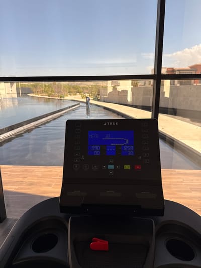 The image displays an indoor perspective from a fitness facility located in San José del Cabo, Mexico, looking out through a large, multi-paned window. In the foreground, the black control console of a "TRUE" brand treadmill is prominent. Its blue digital display shows various metrics, including "METS 33", "0.90", "0.0", "12.58", and "30". A small red safety clip is placed in a recessed storage tray below the console, adjacent to an empty circular cup holder. The interior floor features light-toned wooden planking.
Through the window, an expansive outdoor scene is visible under a clear, bright blue sky with minimal cloud cover. A long, rectilinear water feature, resembling a decorative pool or canal, traverses the midground horizontally, bordered by light-colored concrete pathways. The water surface shows reflections of the sky and surroundings. Within this water feature, a single individual is observed standing, facing away from the camera, and operating a long-handled tool, consistent with cleaning or maintenance. Beyond the water and pathways, the landscape features arid vegetation and light-colored, multi-story buildings with modern architecture, indicative of a resort or residential complex. A building with a reddish-orange tiled roof is partially visible on the far right horizon.