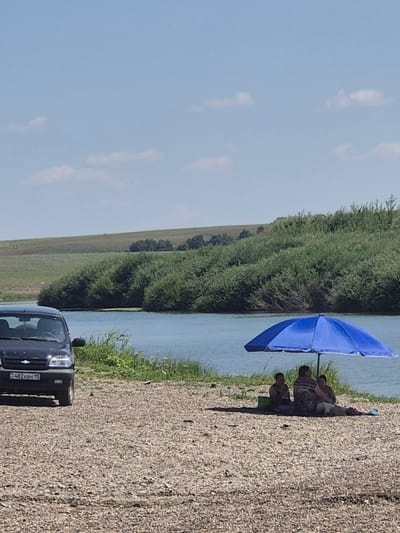 A sunny day at a riverbank near Новоишим ауылдық округі, Kazakhstan, is depicted. A dark-colored SUV is parked on the gravelly shore. In the middle ground, a blue umbrella provides shade for three individuals sitting on a blanket. The river is calm and reflects the blue sky, which is dotted with a few white clouds. Lush green vegetation lines the opposite bank, and rolling hills are visible in the background. The scene suggests a leisurely outing by the water.