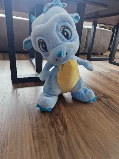 A light blue and yellow plush toy, depicting a stylized dragon or creature, is positioned centrally on a brown wooden or laminate floor in an indoor environment located in Niedernhausen, Germany. The plush toy features prominent round eyes with black pupils and blue irises, a light blue snout, and small, light blue spikes on its head and ear regions. Its torso is light blue, transitioning to a yellow belly. Its feet are light blue with pointed cyan accents. The background reveals the dark, rectangular metal or wooden legs of a table, beneath which a light-colored, upholstered sofa or armchair with vertical stitching details is partially visible. The flooring exhibits visible wood grain patterns and minor surface abrasions. No individuals are present in the scene, and no specific actions or interactions are occurring.