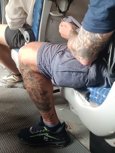 The image shows a close-up view of a person sitting on what appears to be public transportation, possibly a bus, with a distinctive blue and white patterned seat visible. The person is wearing dark blue shorts and black athletic shoes with green and white accents. The person has extensive tattoos on their left leg and arm. The left leg tattoo appears to be a detailed illustration, and there is also text visible on the ankle area. The person's right hand is holding a smartphone. The background shows parts of the interior of the vehicle, including grey flooring and seating components. There is no clear indication of the specific city or location, and therefore no specific details about Kurnool, India can be provided from this image.