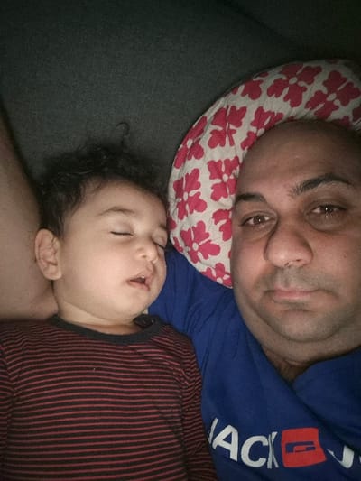 The image displays an adult male and an infant lying together in an indoor setting. The adult male, positioned on the right, is bald, has dark eyes, and is looking towards the camera. He wears a blue t-shirt with a partially visible white and red logo reading "ACK" followed by a red rectangular emblem. A white pillow with a repeating red floral or geometric pattern supports his head. The infant, positioned to the left of the adult, has dark, curly hair and is asleep, with eyes closed and mouth slightly open. The infant wears a long-sleeved shirt with horizontal red and black stripes. Both individuals are situated on a dark grey fabric surface, suggesting a bed or sofa. The ambient lighting indicates an interior environment. The precise location within the city of Melle, Belgium, is not discernible from the visual evidence presented in the image.