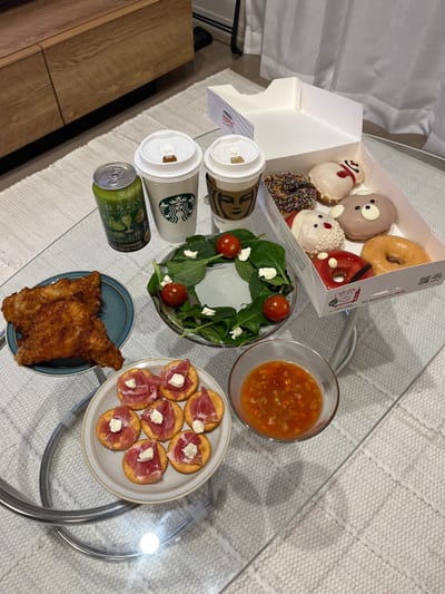 The image displays a spread of food and beverages arranged on a clear glass coffee table over a light-colored, patterned woven rug in a residential setting in Kawasaki, Japan.

In the background, a wooden cabinet is visible to the left, and a light-colored curtain to the right.

The table features two Starbucks-branded white paper cups with brown sleeves and white plastic lids. Adjacent to them is a green aluminum can of Suntory Horoyoi, labeled with Japanese text indicating a 3% alcohol content and grape flavor.

An open white Krispy Kreme doughnut box contains six assorted doughnuts: one with chocolate sprinkles, one white-iced with a red swirl, one brown bear-shaped, one white Santa Claus-themed, one red-iced with black accents, and one plain glazed doughnut. The box features Japanese text.

A small, round, dark teal plate holds three pieces of golden-brown breaded fried chicken. A clear glass bowl contains a salad consisting of green leafy vegetables, several halved cherry tomatoes, and small dollops of white cheese, arranged in a ring.

A beige ceramic plate with a brown rim holds seven round crackers, each topped with a folded slice of cured ham and a small dollop of white soft cheese. A separate small clear glass bowl contains an orange-red chunky mixture, consistent with salsa or a vegetable relish, with visible diced vegetables.

No individuals are present in the frame. The scene is static, depicting a prepared meal or snack assortment.