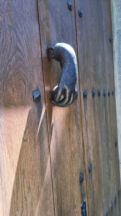 The image shows a close-up of a rustic, weathered wooden door with a unique, cast-iron door knocker shaped like a hand. The door is constructed from vertical planks of dark wood, some showing visible grain and wear. Several metal studs, likely for reinforcement or decoration, are embedded in the wood. Sunlight casts a strong diagonal shadow across the left side of the door, highlighting the texture of the wood. The hand-shaped door knocker, also made of dark metal, is the focal point, with the fingers curled as if holding an invisible object. To the right of the door, a portion of a stone structure is visible. This door knocker is a distinctive feature of the historic town of Rupit i Pruit, Spain, known for its well-preserved medieval architecture. The scene is static and appears to be an exterior shot, likely on a sunny day.