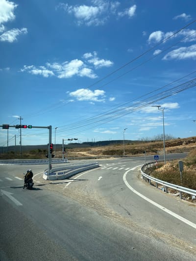 A winding asphalt road curves around a hillside. A white line defines the curve, and a guardrail protects the roadside. On the opposite side, traffic lights stand tall, some displaying red and others green. A motorcyclist waits at the intersection. Overhead, numerous power lines stretch across a bright blue sky filled with scattered white clouds. The arid landscape slopes away from the road, dotted with trees and foliage.