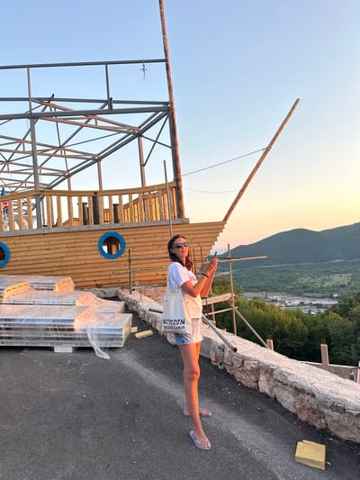In Svinjišta, Montenegro, a woman stands on an asphalt surface, holding a smartphone. She is wearing sunglasses, a white t-shirt with text on it, and denim shorts. To her left, a wooden structure resembling a ship is under construction, with visible metal framing and wooden planks. Several stacks of wrapped construction materials are visible in front of the ship structure. The background shows a lush green landscape with rolling hills under a clear, pale sky.