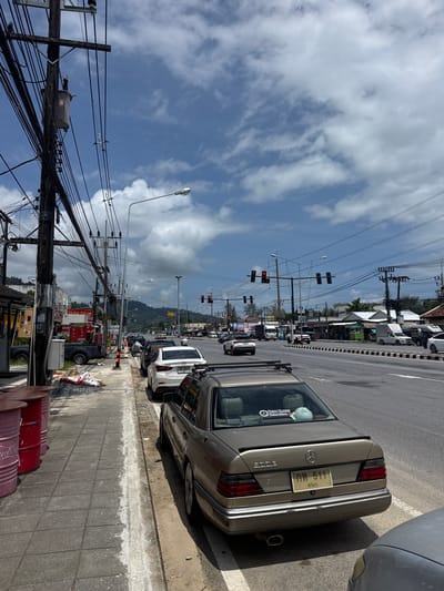 This image shows a street scene in Ban Bang Niang, Thailand, on a cloudy day. The foreground features a gold Mercedes-Benz sedan parked on the side of a multi-lane road. Next to the car, a sidewalk is lined with several large, cylindrical containers, some of which appear to be red metal barrels. Construction materials, including piles of gravel and bags, are also visible on the sidewalk.
The road is moderately busy with various vehicles, including cars, trucks, and motorcycles. Traffic lights are visible overhead, with some showing red signals. On the left side of the road, there are buildings with awnings and signage, suggesting commercial establishments. A prominent wooden utility pole covered in a dense network of electrical wires and a transformer stands near the sidewalk. In the background, a mountain range is visible under a sky filled with white, puffy clouds. The overall impression is that of a typical bustling street in a Thai town.