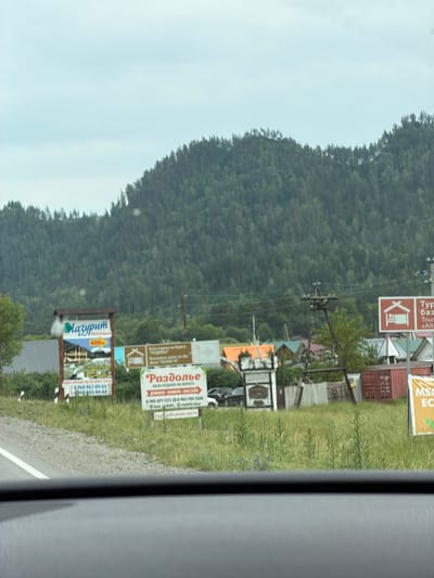 The image shows a roadside view in Cheposh, Russia, with a backdrop of a densely forested mountainside under a cloudy sky. In the foreground, a paved road runs along the left side, transitioning to a gravel shoulder. Several billboards are visible along the right side of the road. One prominent billboard, labeled "Лазурит" (Lazurite), advertises a resort with a price of "400р" (400 rubles) and features an image of a wooden structure. Further down the road, another billboard reads "Раздолье" (Razdolie), indicating a "Recreation center on the shore 'RAZDOLYE'" and listing phone numbers. To the right of this, a billboard with a house icon and the text "Typ" (possibly part of a word like "Tyurma" meaning prison, or a misspelling) and "Alli" (possibly part of "Altai") is visible. The environment is rural, with tall green grass and scattered vegetation. Various structures, including wooden fences, a shipping container, and buildings with pitched roofs, are present behind the billboards. A wooden, industrial-looking structure resembling a historical piece of machinery or a signpost stands in the middle ground. The image appears to be taken from inside a vehicle, as indicated by the dark dashboard visible at the bottom.