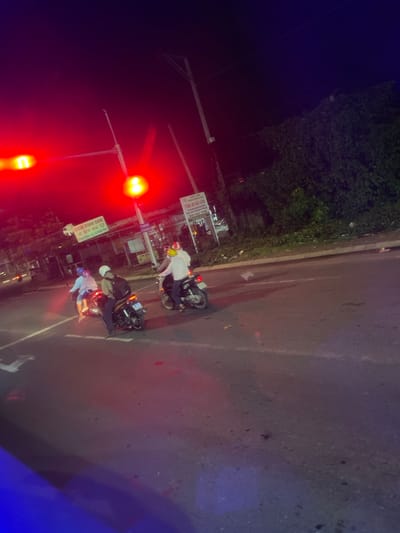 The image shows a nighttime scene on a street in Binh Thuan Ward, Vietnam. Several motorbikes are present, with people riding them. One motorbike in the foreground has a license plate that reads "61F1-181.41". Another motorbike has a rider wearing a yellow helmet and a white long-sleeved shirt. There are also people on a motorbike with a blue helmet and another with a beige helmet and a backpack. Traffic lights are visible, illuminated by red and yellow lights. A sign with Vietnamese text, possibly advertising food, is mounted on a pole. The street appears to be paved, with white lane markings. Vegetation, likely bushes or trees, is visible in the background, and there is a street light pole. The overall impression is of a busy street at night with motorbikes traversing it.
