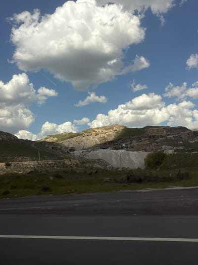 The image shows a landscape in Arzni, Armenia. A mountain with rocky terrain and sparse vegetation is visible. A road runs alongside the mountain, featuring a white line marking the lanes. The sky is blue with scattered white clouds. The scene is captured from a perspective looking slightly upwards towards the mountain and sky.