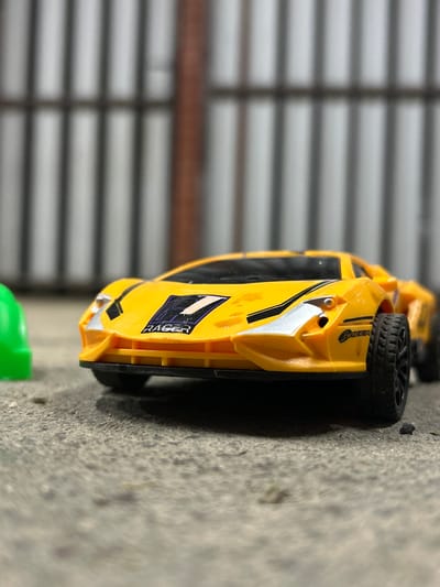 A yellow toy race car is the main subject of this image. The car is positioned on a textured, grey surface, possibly concrete or asphalt, and it is oriented vertically in the frame. The car has black wheels and racing decals on its side, including a large number "7" outlined in purple and white, with the word "RAGER" written in a stylized font on the car's body. There is also a silver accent on the front fender.

In the background, a grey, slatted surface is visible, resembling blinds or a garage door. A thin orange band runs horizontally across this background. To the upper left of the car, a blurred green object can be seen. The lighting suggests it might be daytime, possibly indoors given the background. The image appears to be a close-up shot focusing on the toy car. No people are visible. The overall impression is a simple still life of a toy car. The location context is Dinskaya, Russia.