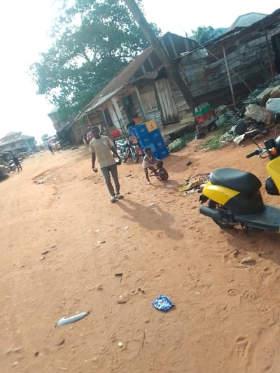The image depicts a dusty, unpaved street scene in what appears to be Orlu, Nigeria. A man wearing a light-colored t-shirt and dark pants walks towards the camera. Two young children are playing near stacks of blue crates and a yellow scooter parked on the side of the road. The street is lined with simple buildings constructed from wood and corrugated metal. Debris and litter are scattered on the ground. Other pedestrians and parked motorcycles are visible further down the street.