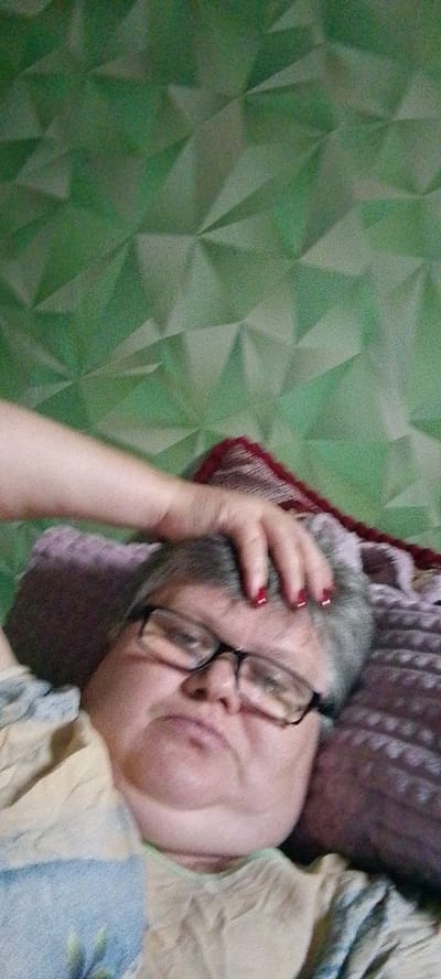 A person with grey hair and glasses is lying down, with their hand resting on their head. The background is a textured green wall. The image appears to be a selfie taken in a bedroom or similar indoor setting. There is no information within the image to indicate the location in Vostochnyi Sosyk, Russia.