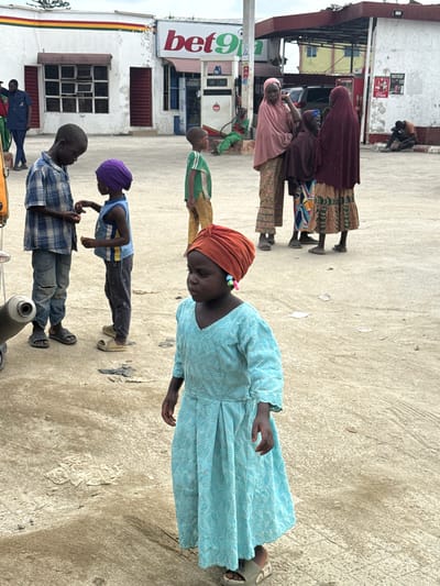 The image depicts a scene at a Bet9ja betting shop in Lagos, Nigeria. In the foreground, a young girl in a light blue dress and orange headscarf stands looking to her right. She wears colorful earrings. Behind her, two boys are interacting. One, wearing a blue and white plaid shirt and torn jeans, appears to be offering something to the other boy, who is in a blue tank top and dark pants, wearing a purple headscarf. Further in the background, other individuals are visible. A man in a blue uniform stands near a building. Three women, two in headscarves and one in a maroon hijab, are conversing. To the right, a man sits on a curb. The environment is an outdoor paved area, possibly a forecourt of a service station, with a building displaying a "Bet9ja" sign. The ground is a mix of concrete and sandy dirt, with some scattered debris. The lighting suggests an overcast day.