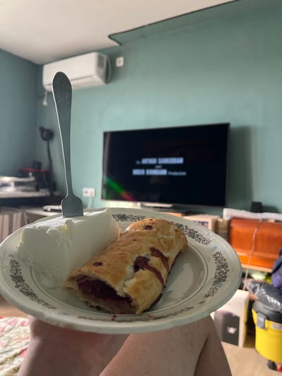 A plate of ice cream and a pastry is held in front of a television screen displaying credits. A fork is inserted into the ice cream. The room appears to be a living room with an air conditioning unit on the wall. The overall scene suggests a person is watching television while eating dessert. The location is not specified.