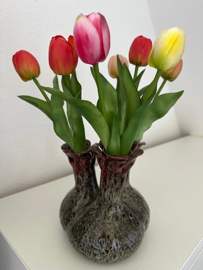 A still life photograph showcases a bouquet of artificial tulips in a dual-chambered, dark grey and red speckled ceramic vase. The vase has a textured, slightly rough surface with a glossy, almost volcanic-looking rim, and it sits on a white surface against a white background. The tulips, with their green stems and leaves, are arranged in varying heights and colors, including pink, red, yellow, and a lighter peach hue. The overall lighting is soft and even, suggesting an indoor setting. The location is not specified in the image itself, and there are no people or discernible environmental features that would indicate a specific city or country like Ossenzijl, Netherlands.