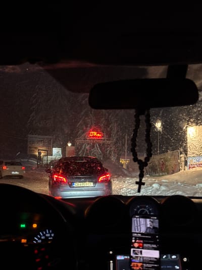 The image depicts the view from inside a car, likely during a winter night in Пампорово, Bulgaria, based on the Cyrillic license plate "CB 6959 CE" on the car ahead and the visible signage with the name "STRE". The windshield is covered with snow. A rearview mirror with rosary beads is in the foreground. The car is in traffic, as indicated by the brake lights of the car in front. The dashboard and instruments of the car are visible on the right side of the frame. A phone is mounted on the dashboard, displaying content on its screen. Signage with text in Cyrillic is visible on the sides of the road. It is snowing.