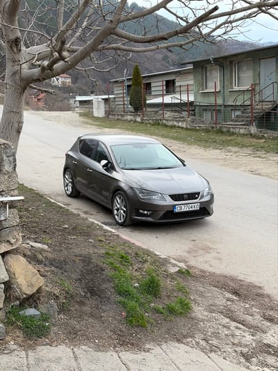 The scene in Tompsan, Bulgaria, depicts a dark grey five-door Seat Leon hatchback, bearing Bulgarian license plate CB 7704 KO, parked on the right side of a dirt and asphalt road. The vehicle is angled slightly towards the upper left of the frame. To the immediate left of the car is a dirt embankment with sparse green grass, bordered by a concrete curb and an old stone and concrete structure. A large, bare-branched deciduous tree stands to the far left. On the opposite side of the road, to the right of the car, are two low-rise buildings; the closer one is a weathered light green structure with concrete steps and red metal railings leading to its entrance. A tall, slender coniferous tree is situated between the buildings and the road. A wire fence runs along the properties. The background features undulating hills or mountains covered with sparse vegetation under an overcast sky. No individuals are visible.