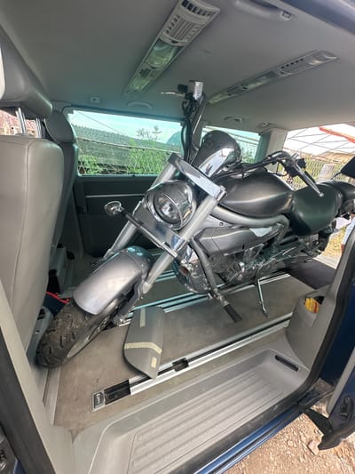 The image shows a motorcycle loaded inside a van. The van appears to be parked outdoors, with a fence and some vegetation visible through the windows. The interior of the van has gray seats and floor mats. The motorcycle is positioned facing forward, with its front wheel secured. The van's side door is open, revealing the exterior ground. The scene suggests an attempt to transport the motorcycle.