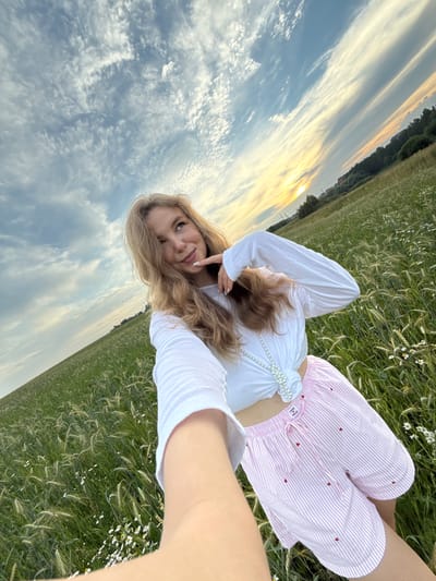 A young Caucasian woman with blonde hair is taking a selfie in a field of tall grass and wildflowers under a cloudy sky at sunset. She is wearing a white long-sleeved shirt tied at the waist and pink and white striped shorts with small red hearts. The field is located in the outskirts of Сновицы, Russia. The sunset casts a warm glow on the horizon, with streaks of orange and yellow clouds. The overall atmosphere is peaceful and serene.