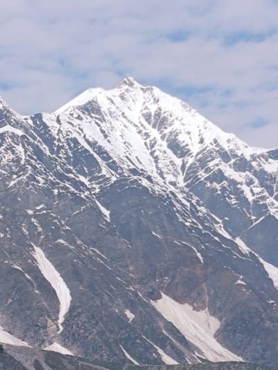 A snow-capped mountain peak dominates the image, with rugged, rocky slopes partially covered in snow. The sky above is a soft blue with scattered white clouds. The mountain's face is etched with the textures of rock and ice, showing streaks of snow running down its steep inclines. The lower portions of the mountain appear to be a darker, more barren rock. The overall impression is one of a vast and formidable natural landscape. The location is identified as Dhundi, India.