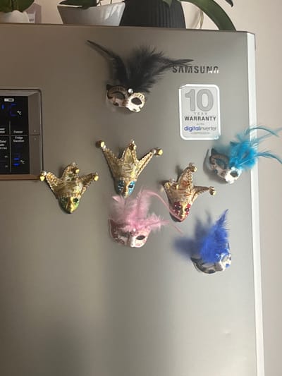 The image shows a close-up of a silver refrigerator door adorned with several Venetian masks and a Samsung warranty sticker. One mask, positioned at the top, is black with feathered accents. Below it, to the left, is a gold-toned mask with green and gold embellishments. To its right, another gold-toned mask with blue accents is visible. Further down, a pink feathered mask with intricate designs is positioned centrally, and a blue feathered mask is seen to its right. The bottom right corner shows a blue feathered mask. The refrigerator also displays a digital control panel on the left side. The background appears to be a domestic indoor setting, with hints of plants and white decorative pots at the top of the frame. There are no people or visible actions taking place. The lighting suggests an indoor environment. The image does not contain any information that places it in Ceraso, Italy.