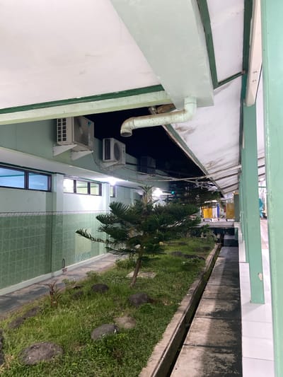This is a landscape photograph taken outdoors, likely in Margoyoso, Indonesia, showing a view down a narrow courtyard or walkway between two buildings. The image is oriented sideways, with the top of the frame on the left.
The left side of the courtyard features the exterior wall of a building, painted a pale green or mint color. A large, white pipe curves downwards along this wall, attached to some outdoor air conditioning units mounted on white brackets. One unit is clearly visible, with a grid-like front. Above these units, the building has a white facade.
The right side of the courtyard is a strip of lush green grass, interspersed with large, dark, round stones that appear to be part of a path or decorative element. A slender tree with needle-like leaves is visible towards the top of this grassy area, casting a shadow. Further down the courtyard, on the right, is a low concrete structure bordering the grass, with what appears to be a drainage channel running alongside it. The wall of the building on this side is made of concrete panels that are weathered and stained.
Further down the courtyard, there are windows visible on the left building, with dark frames and reflections suggesting indoor lighting. The courtyard itself appears to be an outdoor space between buildings, possibly part of a residential complex, school, or other institutional facility. The lighting suggests it might be late afternoon or early evening, with ambient light illuminating the scene. There are no people visible in the image.