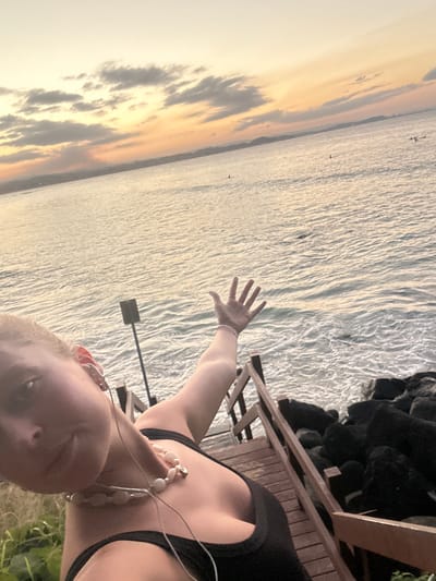 A selfie-style photograph is taken on an elevated wooden deck next to the ocean at sunset. The individual is Caucasian, fair-skinned, and wearing a black top and necklace. A left arm is extended with an open hand toward the ocean. The sea is visible in the background, reflecting the colors of the sunset sky. The horizon has clouds and a golden hue. A black rock outcropping is on the right edge. A ladder is near the deck. The location is suggestive of the coastline in Coolangatta, Australia.