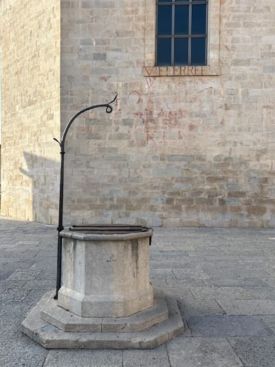 The image shows a stone well in a paved square, with a brick building in the background. The building has a window with multiple panes and some faded red markings on its wall. The well is octagonal in shape and has a metal structure extending upwards and curving over it. There is a shadow cast by a building on the left side of the image onto the well's structure and the wall behind it. The ground is paved with rectangular stones. The scene is an exterior view, likely in an urban setting. This well is located in Plaça de Sant Fèlix in Girona, Spain.