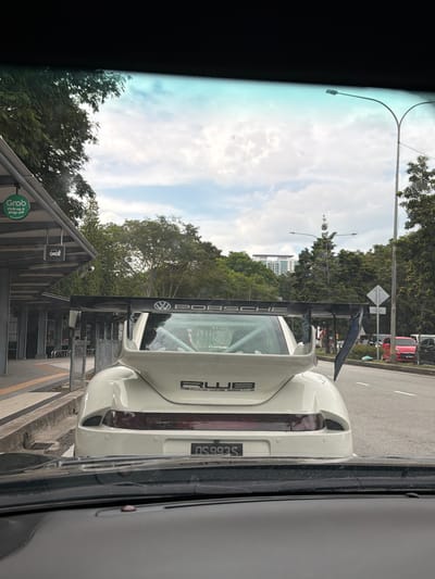 The image depicts the rear of a white RWB (Rauh-Welt Begriff) Porsche 911, identified by its wide body modifications, large black rear spoiler displaying "PORSCHE" and a Volkswagen logo, and a "RWB" badge on the engine cover. A roll cage and "CraftLab" text are visible through the rear window. The vehicle bears a Malaysian license plate, "QS 993 S". The scene is observed from the interior of another vehicle, looking forward through its windshield. To the left of the Porsche is a modern, sheltered pick-up and drop-off point, marked with a green "Grab Pick-up & drop-off" sign and a car icon. A concrete curb separates this area from the asphalt road. The environment includes numerous green trees lining the roadside, a street light pole on the right, and distant high-rise buildings under a partly cloudy sky. Other vehicles are present on the road further down to the right. The scene is situated in Petaling Jaya, Malaysia. No individuals or specific actions are discernible.
