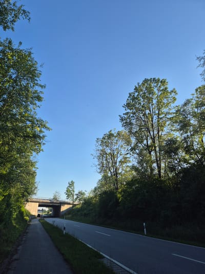 The image captures a sunny day in Essenbach, Germany, with a clear blue sky dominating the upper portion. In the foreground, a paved pathway runs alongside a road. The road itself has white lane markings and is bordered by a grassy verge with white posts. A concrete overpass spans the road and pathway in the middle ground, with trees and foliage on either side. The trees are lush and green, suggesting it is summertime. The scene appears tranquil and undeveloped, with no people or vehicles visible.