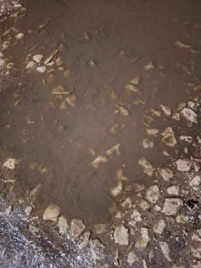 A puddle of murky, brown water fills the lower and central portion of the frame, its surface disturbed by subtle ripples suggesting movement. Scattered throughout the water and along its edges are numerous irregularly shaped fragments of light-colored stone or concrete. The surrounding ground appears to be a mix of wet dirt and gravel. The lighting suggests it is nighttime or a very dark, overcast day. No people, vehicles, or discernible structures are visible. The scene is consistent with a natural accumulation of water and debris on an unpaved surface, possibly a roadside or a disused area. The specific location in Novlyanka, Russia, is not evident from the visual information provided.