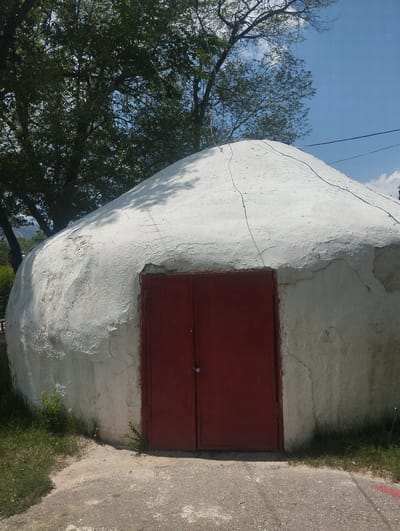 The image shows a white, dome-shaped structure with a red double door in the foreground. The structure appears to be made of concrete and shows signs of wear and tear, with visible cracks and peeling paint. The door is a simple, metallic design with a padlock on the right side. Surrounding the structure is a patch of grass and a paved area. In the background, there are trees and a clear blue sky with some clouds. The scene is likely located in Naryn City, Kyrgyzstan, given the context.