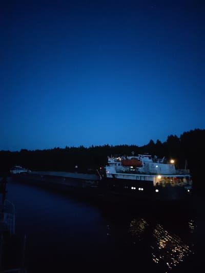 The image depicts a nighttime scene in Volokov Most, Russia, with a clear, dark blue sky overhead, indicating twilight or early night. A vast expanse of water occupies the lower half of the frame, reflecting the lights of the vessels and the distant tree line. A long, dark barge is positioned in the foreground, its structure barely discernible in the dim light. Behind it, a white vessel with illuminated windows and an orange lifeboat is visible. The barge and the vessel appear to be on a river or canal, as evidenced by the calm water. The background is dominated by a dark, dense forest line, silhouetted against the sky, with trees suggesting a natural, possibly rural, setting. The overall atmosphere is serene and quiet.