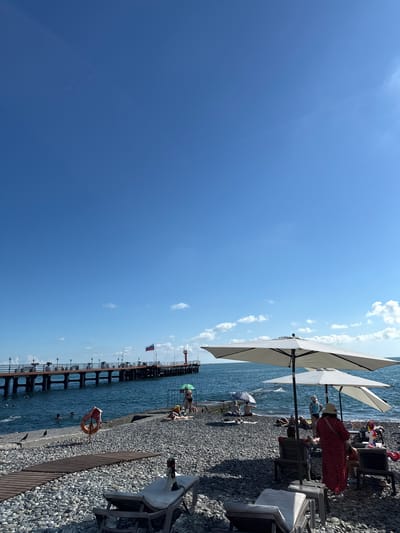 The image depicts a pebble beach on a sunny day with clear blue skies and scattered white clouds. In the foreground, several lounge chairs are positioned on the beach. Two large white umbrellas provide shade for some of the chairs. A wooden walkway leads from the foreground further into the scene.

In the mid-ground, more lounge chairs and people relaxing are visible. Some individuals are sunbathing, while others are seated. A green beach umbrella is open. To the left, in the shallow water near the shore, several people are swimming. A life preserver ring is attached to a pole on the beach.

The background features a long pier extending into the sea, with lampposts lining its length. A Russian flag is visible on the pier, along with a red lighthouse or navigational marker at its end. The sea is a deep blue, with small waves. The location is identified as a beach in Adlerskiy, Russia.