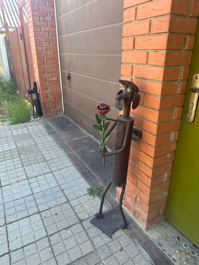 A metal sculpture of a dog holding a red rose stands on a paved area next to a brick pillar and a garage door. The dog sculpture is made of dark metal and has a hat, big eyes, and a snout. The rose appears to be also made of metal, with red petals and a green stem and leaves. The brick pillar is made of orange-red bricks. A brown garage door is visible behind the pillar. To the left of the pillar, a green door is slightly ajar. The ground is paved with square grey tiles. In the background, there is grass and a wooden fence. This scene is located in the city of Temryuk, Russia.