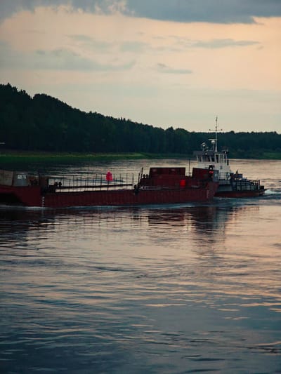 A barge, marked "6076," is being pushed by a tugboat on a wide river. The barge is long and red, with some containers on top. The tugboat is white and grey with a small cabin. A red buoy with the number "3" on it is visible in the water near the barge. The scene is set against a backdrop of a dense, dark green forest lining the riverbank. The sky is overcast with pale clouds, and the water reflects the muted colors of the sky and the surrounding landscape. The overall impression is one of a quiet, working industrial scene in a natural setting. The image was taken in Kazachinskoe, Russia.