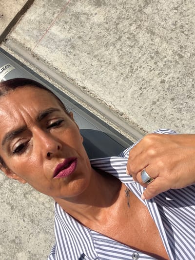 An overhead shot shows a woman in Roodt-sur-Syre, Luxembourg, with her face tilted towards the sky. She is wearing a striped shirt and a silver ring on her finger, which is resting on her chest. Her makeup includes dark eyeliner and plum lipstick. There is a small, delicate tattoo of a branch on her collarbone. The background is a concrete surface, possibly a wall or pavement. The lighting suggests it is a sunny day.
