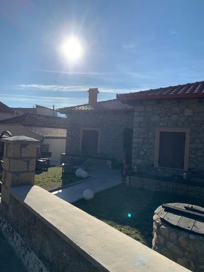 The vertically oriented image depicts a bright, sunny outdoor scene in Kastraki, Greece, showcasing traditional stone architecture under a clear blue sky.
The main subject is a stone house with a terracotta-tiled roof, featuring at least two windows with dark wooden shutters. A chimney is visible on its roofline. Another building with a similar tiled roof is partially seen behind the main house.
The sky, dominating the upper and left portions of the image, is a vivid blue with a bright, intense sun positioned in the lower-left, creating a significant lens flare. Several faint white contrails are visible against the blue expanse.
The ground area around the house consists of green grass and a light-colored paved pathway. Two modern spherical white outdoor lights are placed along this path. To the right, a rustic well-like structure or decorative wooden barrel, crafted from stone and wood, stands on the grass. A robust, light-colored concrete or stone wall extends diagonally across the foreground on the right side.
The scene conveys a sense of clear weather and bright daylight, likely during the morning or afternoon. The overall atmosphere is serene and inviting. No discernible text is visible within the image.