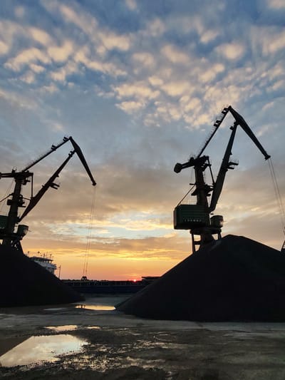 This image captures a port scene at sunset, featuring two large industrial cranes silhouetted against a dramatic sky. The cranes are positioned on either side of the frame, with their long arms reaching upwards and outwards. Piles of dark material, likely coal or ore, are visible at the base of each crane. In the foreground, puddles of water reflect the warm hues of the sunset. A ship is partially visible in the background to the left, and a long structure, possibly a pier or loading dock, stretches across the horizon. The sky is filled with scattered clouds tinged with orange and yellow from the setting sun, creating a striking contrast with the dark silhouettes of the cranes and the industrial landscape. The location is identified as Chelganovo, Russia.