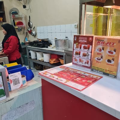 The image depicts an eatery counter with advertisements. One is a "PROMO JULEHA," and another advertises "Diskon KILAT" (Flash Discount). A sign mentions "AYAM LEBIH BANYAK..." (More Chicken) with a price range, indicating a deal on chicken. It states "HARGA LEBIH MURAH" (Cheaper Price) at Rp. 20,000. The area includes food preparation items, shelving, and a person wearing a headscarf. QR codes are visible.