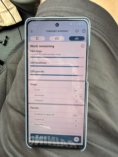 The image shows a smartphone displaying an itinerary summary. The screen shows information about work remaining, including the number of stops, locations, and parcels. It also details the status of stops and parcels, categorizing them into "To Do", "Successful", and "Problems". The current stop is indicated at the bottom of the screen. The phone is held by a person wearing dark fabric clothing. The background is out of focus, but appears to be an indoor environment with some textured surfaces. There is no visible indication of the city of Shifnal, United Kingdom.